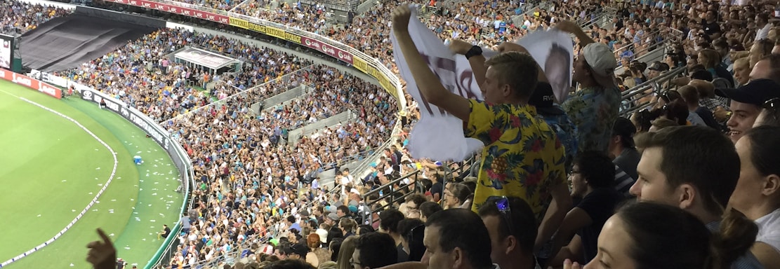 A crowded stadium filled with spectators during a sports event. The audience is animated, with some individuals holding signs and others clapping. The stadium is well-lit with a clear view of the field on the left side. The architecture of the stadium has a modern, open-roof design.