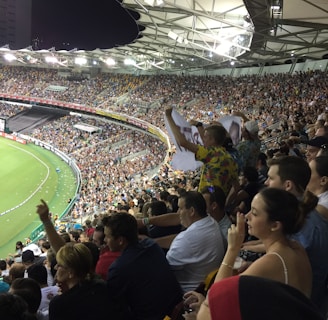 A crowded stadium filled with spectators during a sports event. The audience is animated, with some individuals holding signs and others clapping. The stadium is well-lit with a clear view of the field on the left side. The architecture of the stadium has a modern, open-roof design.