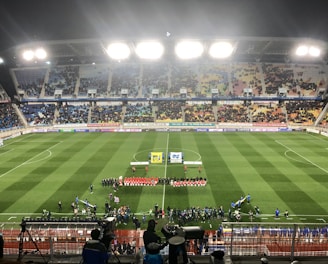 A large soccer stadium with bright overhead lights and a well-maintained green field. Teams are lined up in the center alongside officials, and there is a group gathered nearby, possibly for a pre-match ceremony. The stadium is filled with spectators wearing colorful clothing, and banners and advertisements are visible along the edges of the field. Cameras and officials are positioned on the sidelines to capture the event.