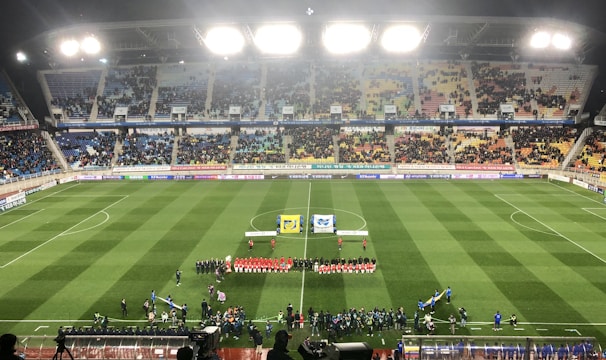 A large soccer stadium with bright overhead lights and a well-maintained green field. Teams are lined up in the center alongside officials, and there is a group gathered nearby, possibly for a pre-match ceremony. The stadium is filled with spectators wearing colorful clothing, and banners and advertisements are visible along the edges of the field. Cameras and officials are positioned on the sidelines to capture the event.