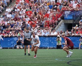 A sports event with two athletes engaged in a lacrosse game. One athlete in a white jersey is holding a lacrosse stick, appearing to prepare for a play. A referee in a striped uniform is nearby. The background features a large audience seated in a stadium, with many spectators wearing bright and casual clothing. Some photographers are also visible near the field.
