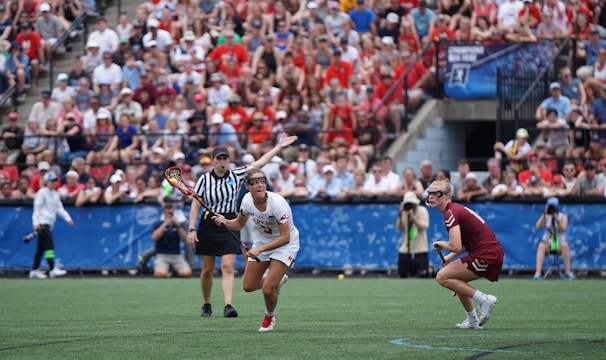 A sports event with two athletes engaged in a lacrosse game. One athlete in a white jersey is holding a lacrosse stick, appearing to prepare for a play. A referee in a striped uniform is nearby. The background features a large audience seated in a stadium, with many spectators wearing bright and casual clothing. Some photographers are also visible near the field.