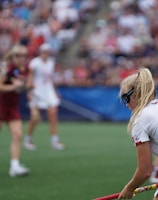 A sporting event with athletes on a grass field, one in a white jersey with the number 11 in the foreground, holding a lacrosse stick. Players in the background wear maroon uniforms. Spectators fill the stands creating a lively atmosphere.