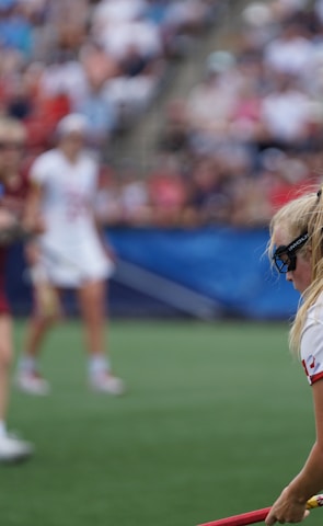 A sporting event with athletes on a grass field, one in a white jersey with the number 11 in the foreground, holding a lacrosse stick. Players in the background wear maroon uniforms. Spectators fill the stands creating a lively atmosphere.