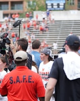 A lively gathering in an outdoor sports venue with a group of people, some wearing matching shirts labeled 'National Champions.' Several individuals are holding cameras and microphones, indicating a media presence. The background shows blurred spectators sitting on bleachers.