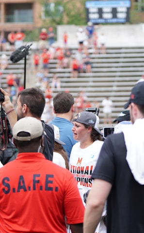A lively gathering in an outdoor sports venue with a group of people, some wearing matching shirts labeled 'National Champions.' Several individuals are holding cameras and microphones, indicating a media presence. The background shows blurred spectators sitting on bleachers.