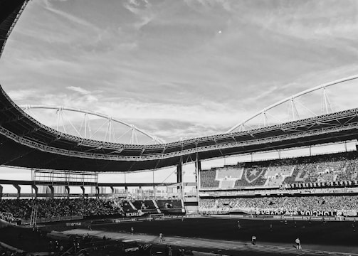 A large sports stadium filled with spectators. The stadium features a curved roof structure and seats filled with people. The sky is partially cloudy, and the field is prepared for an event.