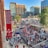 A vibrant outdoor scene featuring a large crowd of people gathered near a sports venue. A prominent red banner with '2018' and a sports logo hangs from a brick building. Nearby, there is a sign for a team store and souvenir shop. Surrounding the event are several modern high-rise buildings and a parking lot full of cars. Tall trees are visible in the background.