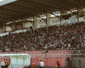 A crowded stadium with spectators filling the stands, observing an event. The stadium is covered and has multiple sections labeled with various advertisements visible on the walls. In front of the stands, a few people are gathered near an area with chairs and a white canopy.