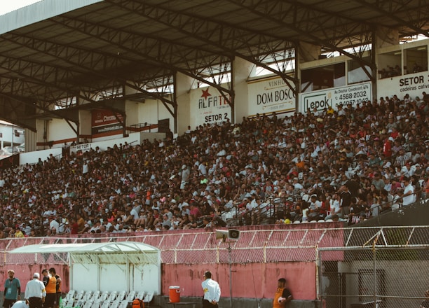 A crowded stadium with spectators filling the stands, observing an event. The stadium is covered and has multiple sections labeled with various advertisements visible on the walls. In front of the stands, a few people are gathered near an area with chairs and a white canopy.