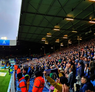 A crowded stadium filled with enthusiastic spectators. Many are standing and appear to be watching a sporting event with intensity. The stadium lighting is on, and the field is partially visible to the left. Some people are wearing team apparel, suggesting a sports team atmosphere. There are stewards wearing bright orange jackets along the edge of the field.