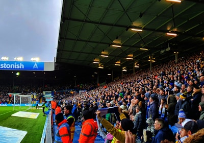 A crowded stadium filled with enthusiastic spectators. Many are standing and appear to be watching a sporting event with intensity. The stadium lighting is on, and the field is partially visible to the left. Some people are wearing team apparel, suggesting a sports team atmosphere. There are stewards wearing bright orange jackets along the edge of the field.