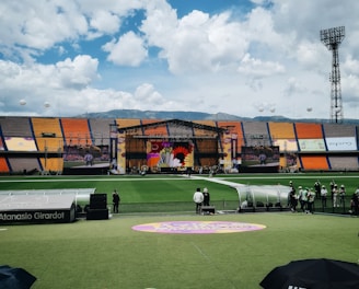 A sports stadium with a large stage set up on the field, hosting an outdoor event. Spectators wearing colorful hats sit in the foreground. The seating areas display vibrant colors, and a clear blue sky with scattered clouds is visible. Large screens display graphics related to the event.