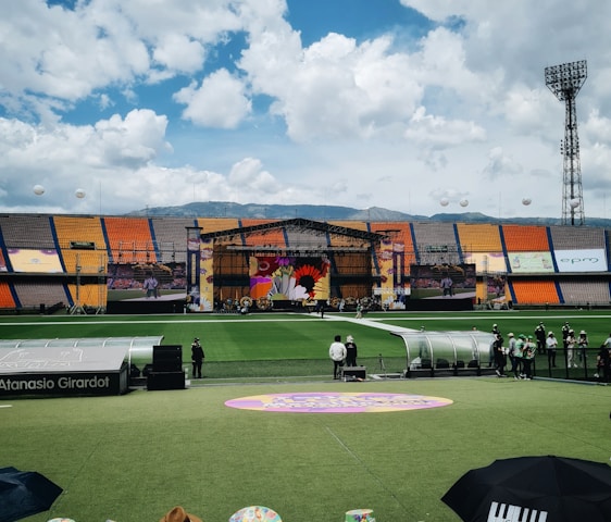 A sports stadium with a large stage set up on the field, hosting an outdoor event. Spectators wearing colorful hats sit in the foreground. The seating areas display vibrant colors, and a clear blue sky with scattered clouds is visible. Large screens display graphics related to the event.