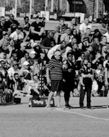 A large crowd of people is gathered, with some standing and others sitting on bleachers. Several individuals are wearing sports attire, and there is a field visible in the foreground with a marker indicating 50 meters. The scene appears to be a sporting event, with a diverse group of spectators.