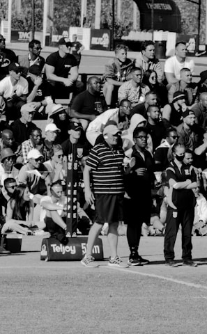 A large crowd of people is gathered, with some standing and others sitting on bleachers. Several individuals are wearing sports attire, and there is a field visible in the foreground with a marker indicating 50 meters. The scene appears to be a sporting event, with a diverse group of spectators.