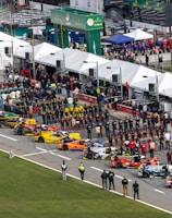 Aerial view of a motorsport event featuring a grid of colorful racing cars lined up in a row. Many people, including teams in uniforms and spectators, are gathered alongside tents and pit lane structures. The scene is bustling with activity, and there are various promotional signs and banners visible.