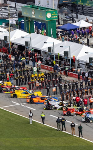 Aerial view of a motorsport event featuring a grid of colorful racing cars lined up in a row. Many people, including teams in uniforms and spectators, are gathered alongside tents and pit lane structures. The scene is bustling with activity, and there are various promotional signs and banners visible.
