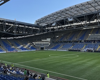 A modern sports stadium with a large, green soccer field and surrounding seating areas. Blue and yellow seats are visible, with some spectators scattered across the stands. The stadium is partially covered with a steel lattice roof, showing a clear blue sky. Several security personnel in high-visibility vests are present.
