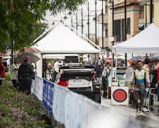 A bustling rally service park featuring race cars and teams preparing for an event. Several individuals are gathered under tents, dressed in racing suits and team jackets. The setting appears lively and organized amidst a light rain, with spectators holding umbrellas and observing the preparations.