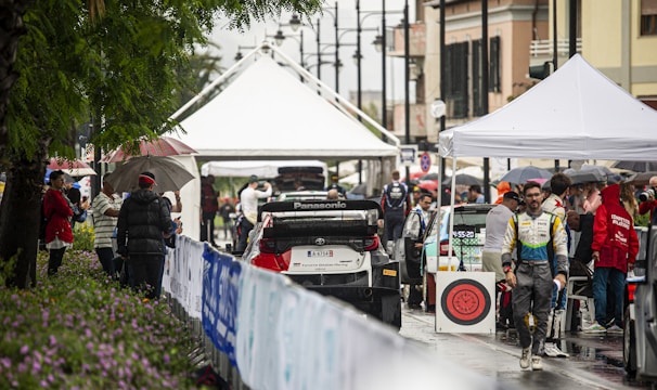A bustling rally service park featuring race cars and teams preparing for an event. Several individuals are gathered under tents, dressed in racing suits and team jackets. The setting appears lively and organized amidst a light rain, with spectators holding umbrellas and observing the preparations.