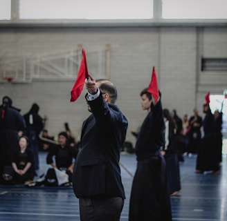 A martial arts event taking place in an indoor sports hall. Several individuals dressed in traditional martial arts attire participate, some wearing protective gear. A person prominently holds up a red flag, potentially indicating a referee or judge in action. The background includes spectators and sports equipment.
