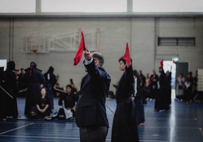 A martial arts event taking place in an indoor sports hall. Several individuals dressed in traditional martial arts attire participate, some wearing protective gear. A person prominently holds up a red flag, potentially indicating a referee or judge in action. The background includes spectators and sports equipment.