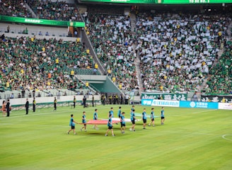 A large sports stadium is filled with spectators, the majority wearing green shirts, creating a vibrant and energetic atmosphere. In the foreground, a group of individuals dressed in blue shirts are carrying a large flag across the field. Stadium staff and officials are positioned near the field and among the crowd.