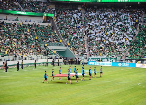 A large sports stadium is filled with spectators, the majority wearing green shirts, creating a vibrant and energetic atmosphere. In the foreground, a group of individuals dressed in blue shirts are carrying a large flag across the field. Stadium staff and officials are positioned near the field and among the crowd.