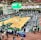 A large indoor arena with a basketball court at the center, surrounded by multiple stalls set up for a graduate and professional schools fair. The area is bustling with people walking around, interacting, and gathering information at various booths. A prominent digital screen displays information about the event. Overhead, banners are visible, indicating past sports events.