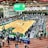 A large indoor arena with a basketball court at the center, surrounded by multiple stalls set up for a graduate and professional schools fair. The area is bustling with people walking around, interacting, and gathering information at various booths. A prominent digital screen displays information about the event. Overhead, banners are visible, indicating past sports events.